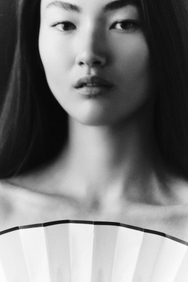 Frontal black and white beauty portrait of a woman with long dark hair holding a white paper fan at her collarbone