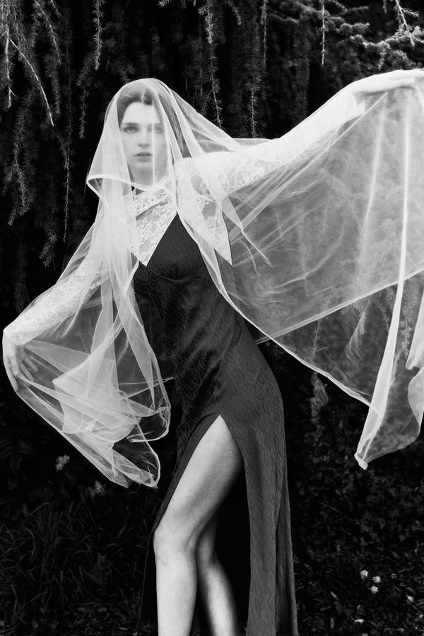 Woman in a black dress and flowing white tulle veil standing in a dark forest, black and white fashion photograph