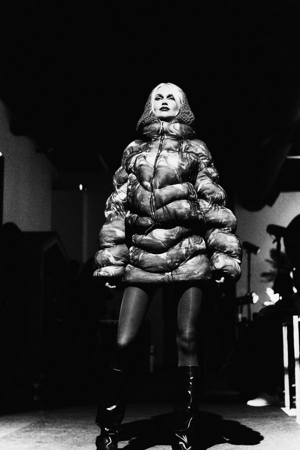 Model in a voluminous fur coat standing in a dim studio between light stands, black and white fashion photograph