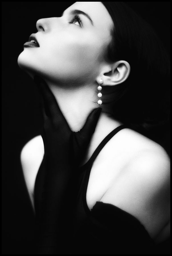 Black and white portrait of a woman in a black top with a pearl drop earring, head tilted back and looking upward against a dark background