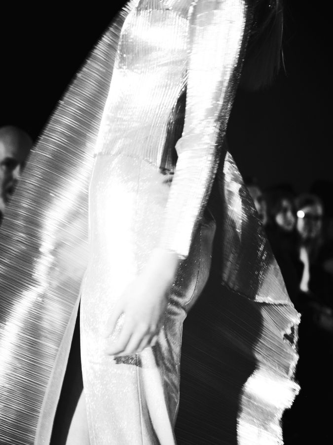 Black and white runway photograph of a model in a metallic pleated gown mid-stride